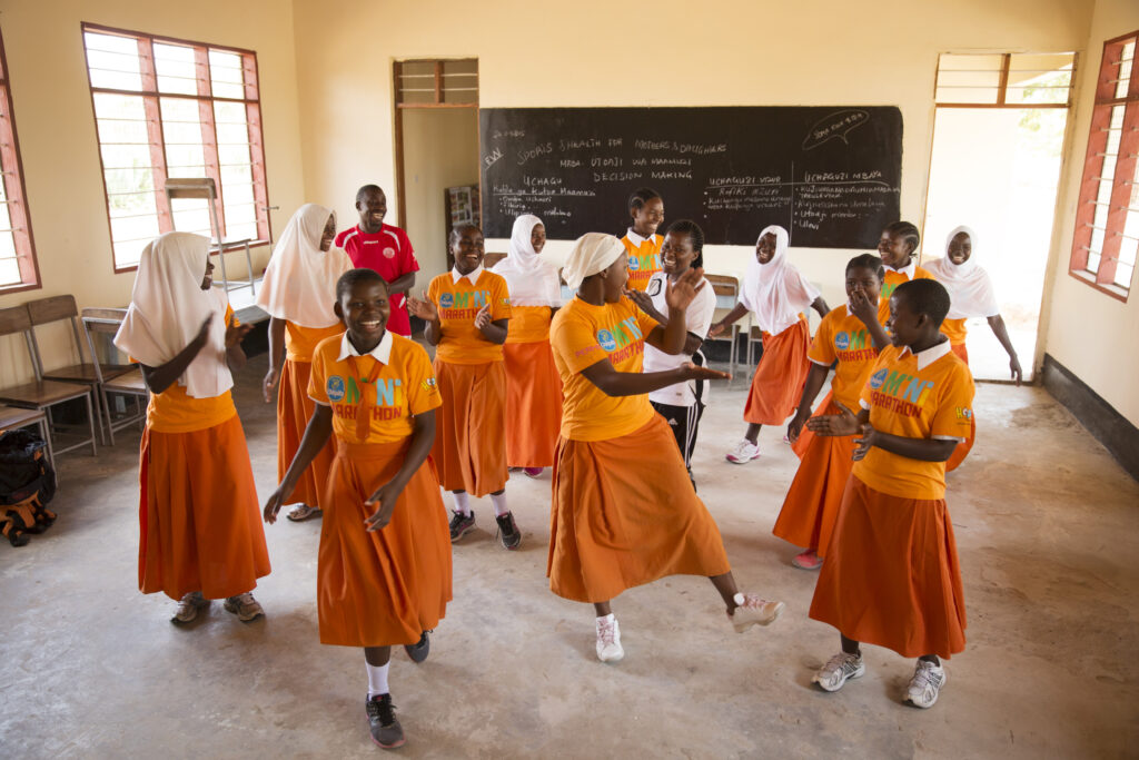 Kuvituskuva tansanialaisen koulun luokkahuoneesta. Kuvassa 12 tyttöä seisoo luokan keskellä tanssimassa. 